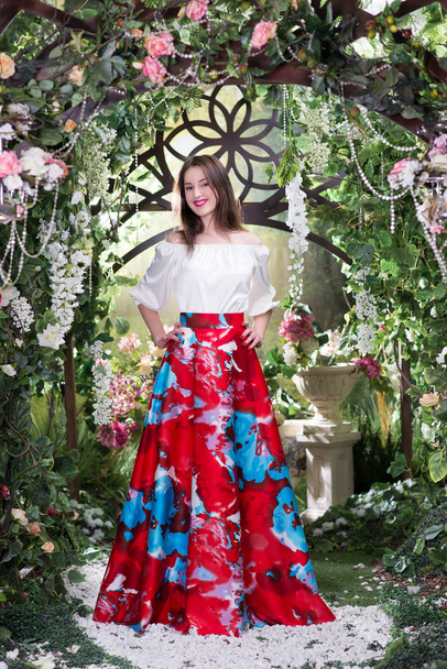 Молода стильна жінка в розкішній червоній спідниці з квітковим візерунком і шовковою блузою позує під квітучою аркою
 - Фото, зображення