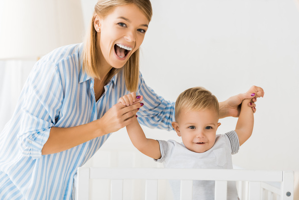cheerful mother looking at camera and holding smiling toddler in hands - Foto, Bild