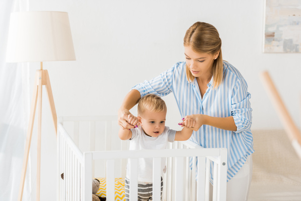 mom holding hands of son in crib in nursery room - Zdjęcie, obraz