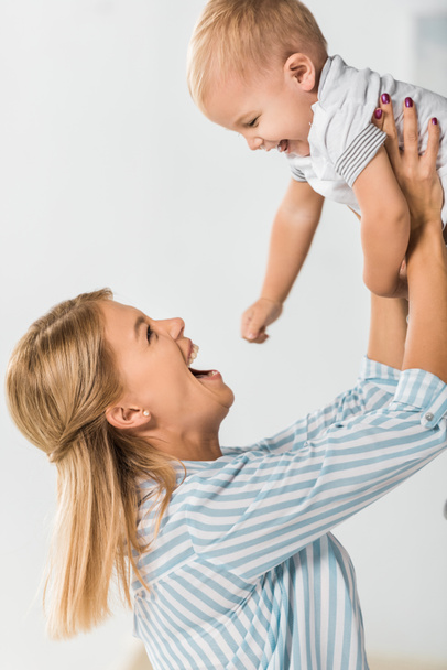 cheerful mother looking at toddler and holding him in hands on white background - Foto, Imagem