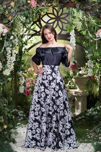Młoda kobieta elegancka w luksusowych czarna sukienka w kwiatowy ornament pozowanie pod łuk kwitnący - Zdjęcie, obraz