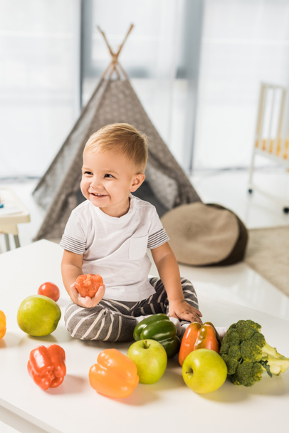 милый улыбающийся ребенок сидит на столе в окружении фруктов и овощей
 - Фото, изображение