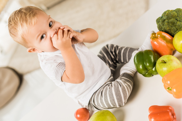cute toddler eating and sitting on table surrounded by fruit and vegetables - Fotografie, Obrázek
