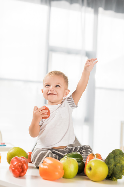 bonito criança levantando a mão e sentado na mesa cercada por frutas e legumes
  - Foto, Imagem