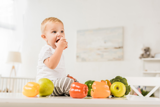 cute toddler eating and sitting on table surrounded by fruit and vegetables - Foto, Bild