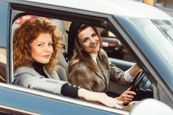 selective focus of smiling stylish woman in black hat driving car while her ginger female friend sitting near  - Zdjęcie, obraz