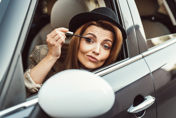 beautiful woman in black hat looking at side mirror while using ink for eyelashes in car  - Zdjęcie, obraz
