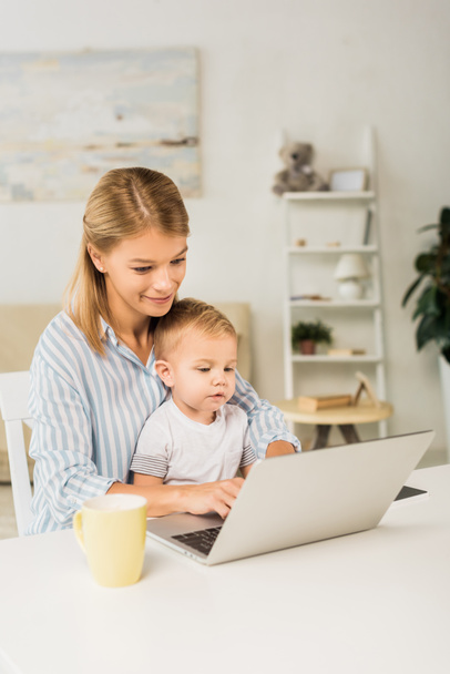 madre sentada en el escritorio con un niño lindo mientras usa la computadora portátil
 - Foto, imagen