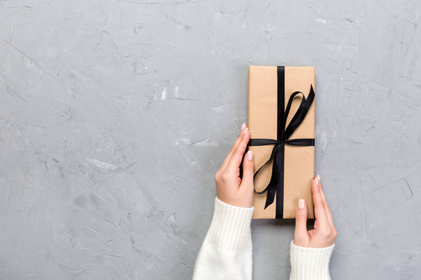 Woman hands give wrapped valentine or other holiday handmade present in paper with black ribbon. Present box, decoration of gift on white cement table, top view with copy space. - Foto, Bild