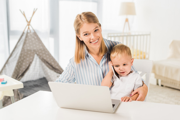 cute toddler holding smaptphone with mother sitting at desk while using laptop - Фото, зображення