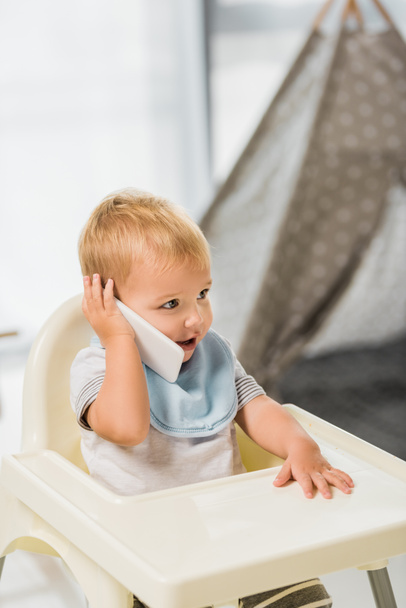 lindo niño hablando en el teléfono inteligente y sentado en silla de bebé
 - Foto, Imagen