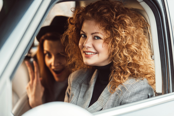 Portret van vrolijke gember vrouw kijken camera terwijl haar vriendin overwinning teken in auto tonen - Foto, afbeelding