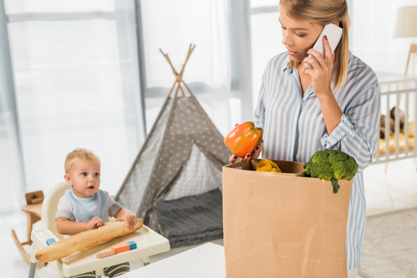 busy mother holding groceries while talking on smartphone with toddler in baby chair on background - Photo, Image