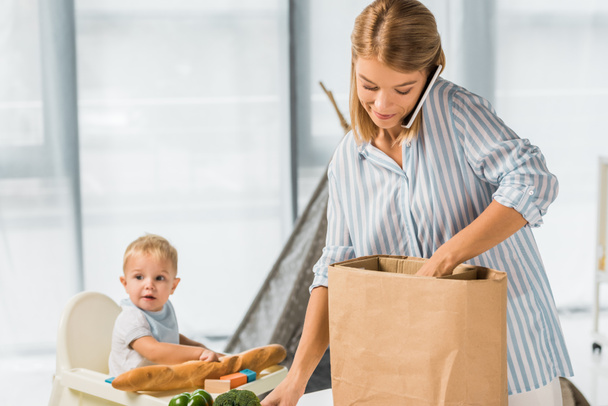 mother holding groceries while talking on smartphone with toddler in baby chair on background - Photo, Image