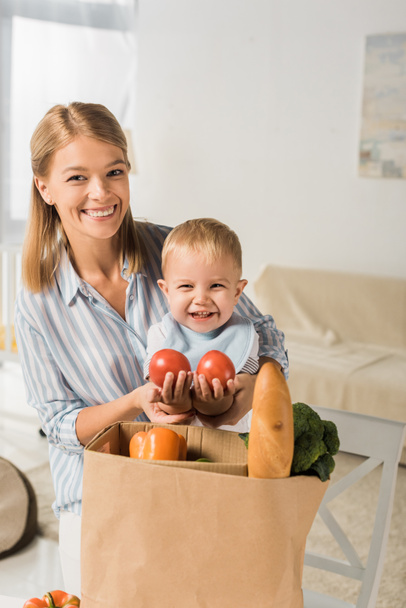 happy mom showing groceries to cheerful son and looking at camera - Фото, зображення