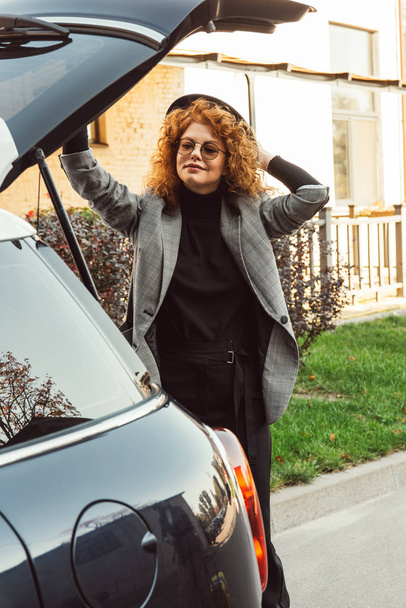 stylish curly redhead woman in grey jacket closing car trunk at city street - Fotografie, Obrázek