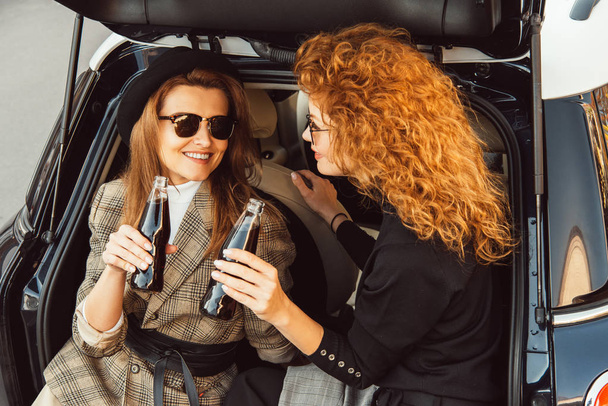smiling adult women in jackets clinking by soda bottles in car trunk at urban street - Zdjęcie, obraz