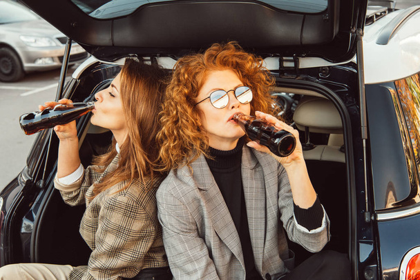 high angle view of female friends in jackets drinking soda in car trunk at urban street - Zdjęcie, obraz