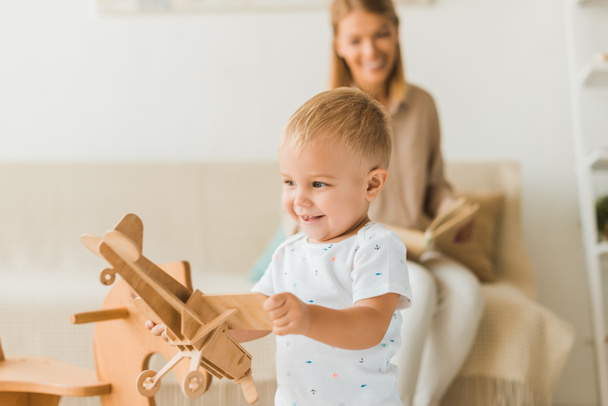 niño feliz jugando con juguete avión de juguete de madera en la habitación del vivero con la madre en el fondo
 - Foto, imagen