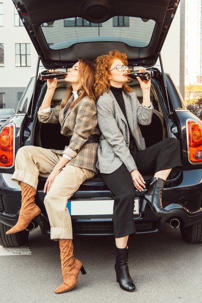 attractive adult women in jackets drinking soda in car trunk at urban street - Fotó, kép