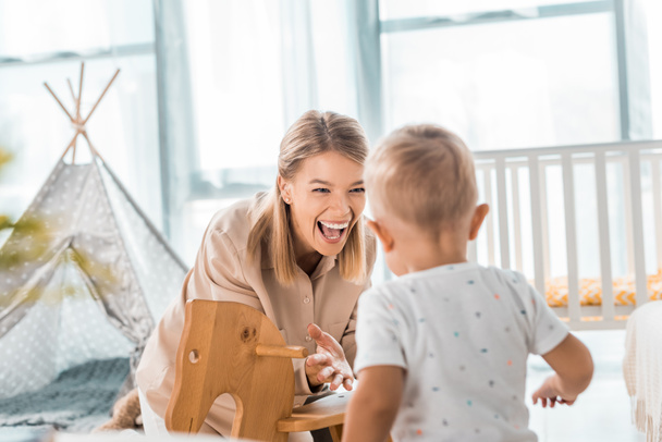 happy mother and toddler playing with toy wooden rocking horse chair in nursery room - Photo, Image