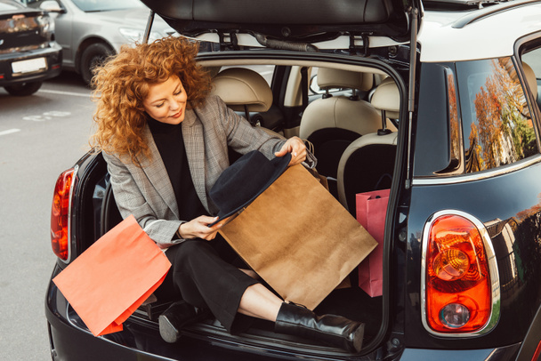 stylish redhead curly woman with black hat and shopping bags sitting in car trunk - Photo, Image