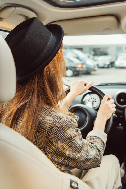 задний вид женщины в черной шляпе и клетчатой куртке, сидящей за рулем в машине
 - Фото, изображение