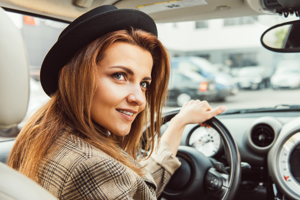 stylish woman in black hat and checkered jacket sitting at steering wheel in car - Фото, зображення