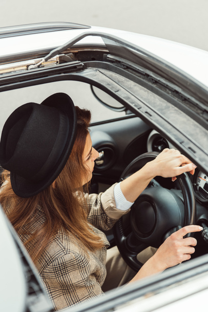 high angle view of fashionable woman in black hat and checkered jacket sitting at steering wheel in car - Φωτογραφία, εικόνα