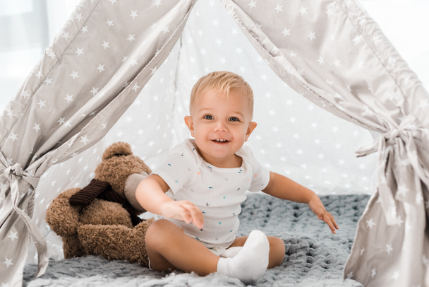 smiling adorable toddler sitting in baby wigwam with fluffy teddy bear toy - Foto, afbeelding