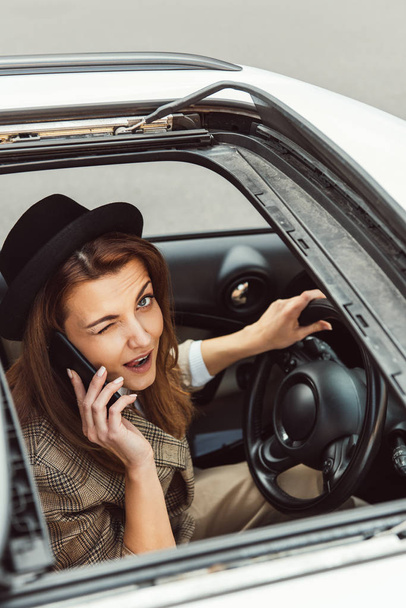 femme heureuse en chapeau noir clin d'oeil et parler sur smartphone tout en étant assis au volant dans la voiture
 - Photo, image