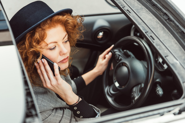 selective focus of beautiful ginger woman in black hat talking on smartphone while sitting at steering wheel in car  - Fotó, kép