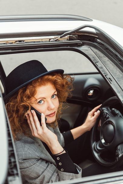 high angle view of attractive ginger woman in black hat talking on smartphone while sitting at steering wheel in car  - Photo, Image