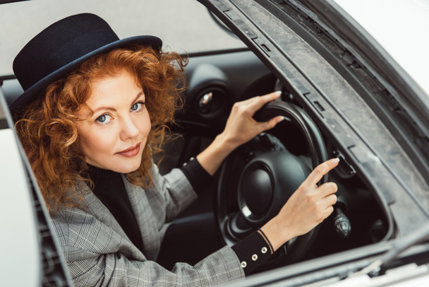 portrait of stylish ginger woman in black hat and grey jacket sitting at steering wheel in car  - Photo, Image