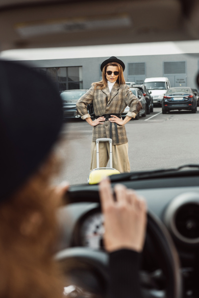 imagem cortada de mulher sentada no volante enquanto turista elegante feminino de pé com as mãos na cintura perto de saco de rodas na rua da cidade
  - Foto, Imagem