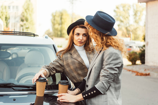 amigas de moda en sombreros negros y chaquetas de pie con tazas de café de papel y mirándose el uno al otro cerca del coche en la calle
 - Foto, Imagen