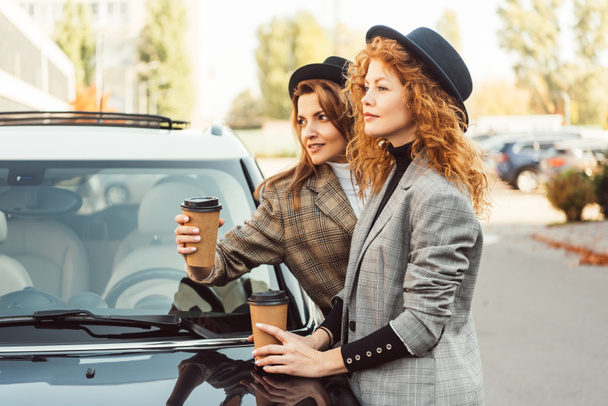 side view of stylish women in black hats and jackets standing with paper coffee cups near car at street - Foto, Imagen