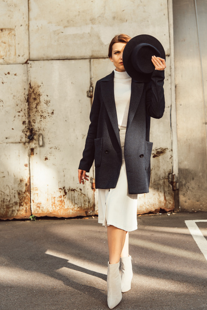 modello femminile adulto in cappotto fumare sigaretta e che copre la faccia da cappello nero in strada urbana
 - Foto, immagini