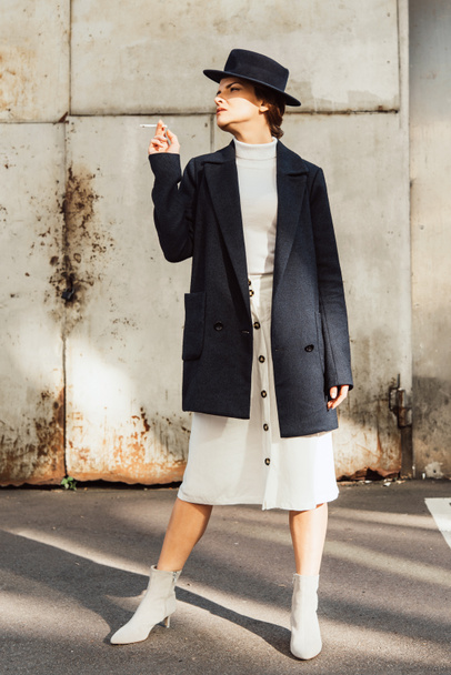 élégant modèle féminin en manteau et chapeau noir posant avec cigarette à la main dans la rue urbaine
 - Photo, image