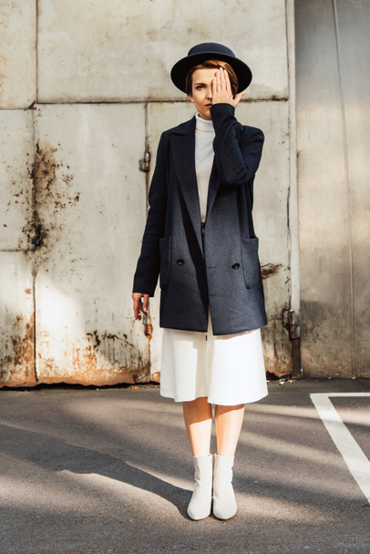 modieuze vrouwelijke model in jas en zwarte hoed poseren met sigaret in de hand bij urban street - Foto, afbeelding