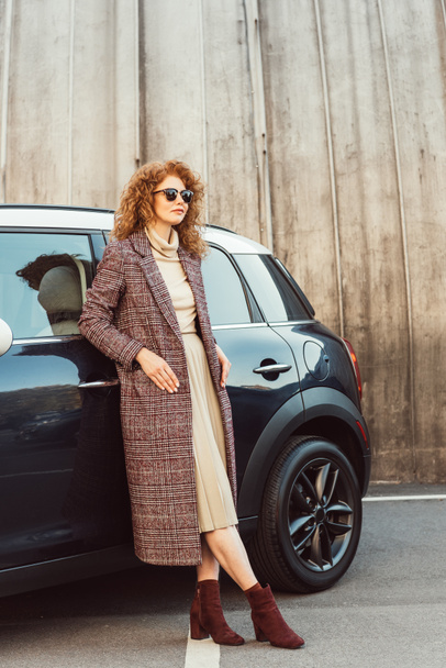 χαμογελώντας κομψή γυναίκα τζίντζερ σε παλτό και γυαλιά ηλίου που παρουσιάζουν κοντά στο αυτοκίνητο στο δρόμο της πόλης  - Φωτογραφία, εικόνα