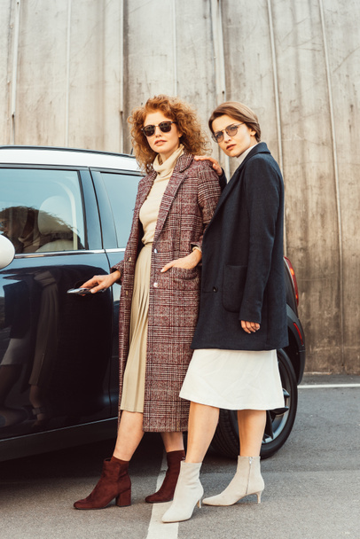 smiling fashionable women in coats and sunglasses posing near car at city street - Photo, Image