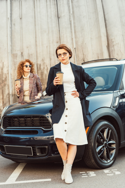 fashionable female models in coats posing with disposable coffee cups near car at urban street - Photo, Image