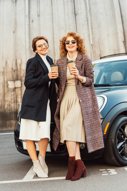 riéndose amigas en abrigos bebiendo café cerca del coche en la calle urbana
 - Foto, Imagen