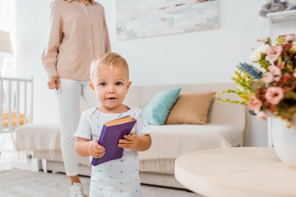 adorable toddler holding book and looking at camera indoors with mother on background  - 写真・画像