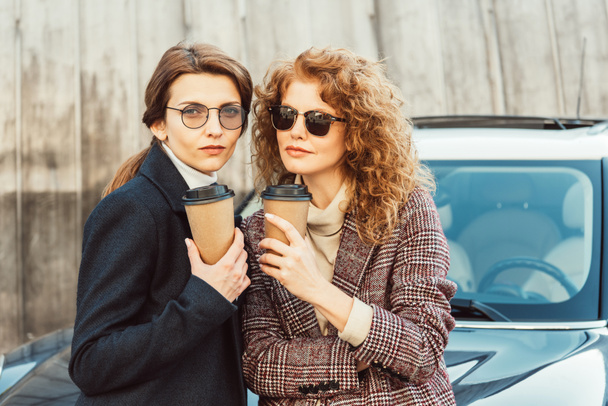 enfoque selectivo de las amigas de moda en gafas de sol y abrigos beber café cerca de coche en la calle urbana
 - Foto, Imagen