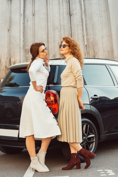 stylish adult female models in sunglasses and skirts posing near car at urban street - Photo, Image