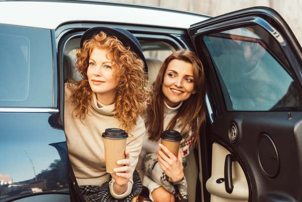 heureux amis féminins élégants avec tasses à café jetables regardant de la voiture à la rue
 - Photo, image