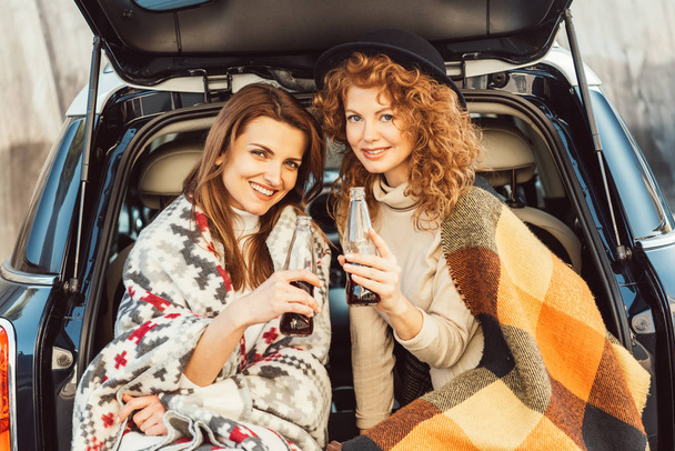 selective focus of happy women wrapped in blankets holding soda bottles sitting in car trunk at urban street - Photo, image