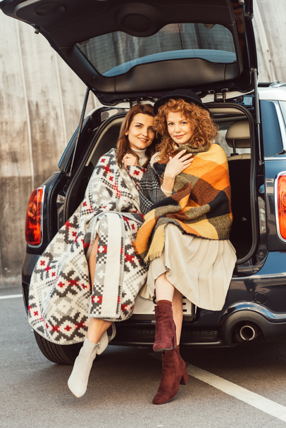 стильные подружки завернутые в одеяла, смотрящие в камеру и сидящие в багажнике машины на городской улице
 - Фото, изображение